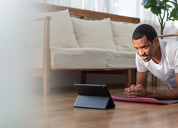 Schwarzer Mann macht eine Sportübung auf der Isomatte in seinem Wohnzimmer. Vor ihm steht ein Tablet. 