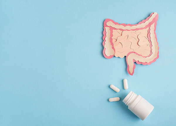 Flatlay: Scherenschnitt des Dick- und Dünndarms, darunter eine Dose, aus der drei Kapseln herausschauen.