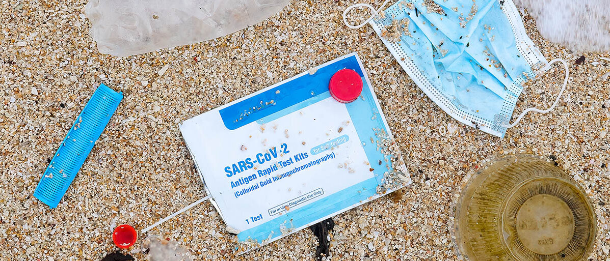 Die Verpackung eines Corona-Schnelltests und eine Maske liegen zwischen anderem Plastikmüll am Strand im Sand; eine Welle spült Schaum heran.