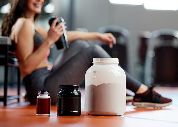 Nahrungsergänzungsmittel: Für wen sind welche Nährstoffe als Nahrungsergänzung wirklich sinnvoll? Eine Frau sitzt im Fitnesstudio auf dem Boden und trinkt einen Shake. Im Vordergrund stehen drei Packungen mit Pulver und Kapseln.