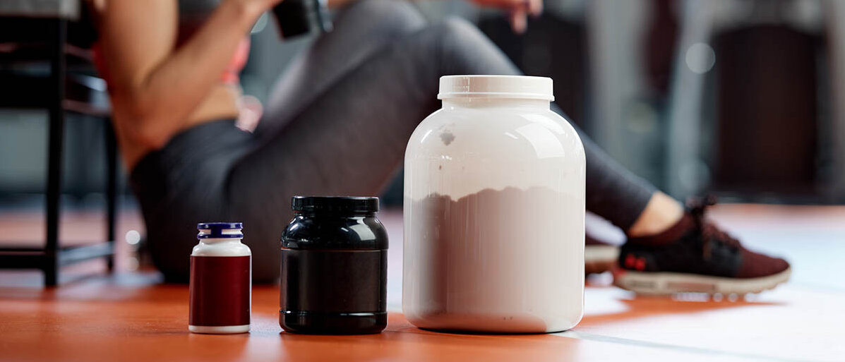Eine Frau sitzt im Fitnesstudio auf dem Boden und trinkt einen Shake. Im Vordergrund stehen drei Packungen mit Pulver und Kapseln.