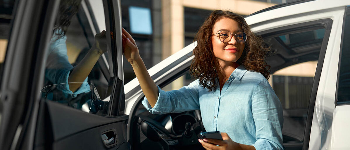 Eine junge Frau steigt aus der Fahrertür eines Autos. Sie trägt eine Bluse, hat ihr Smartphone in der Hand und lächelt.