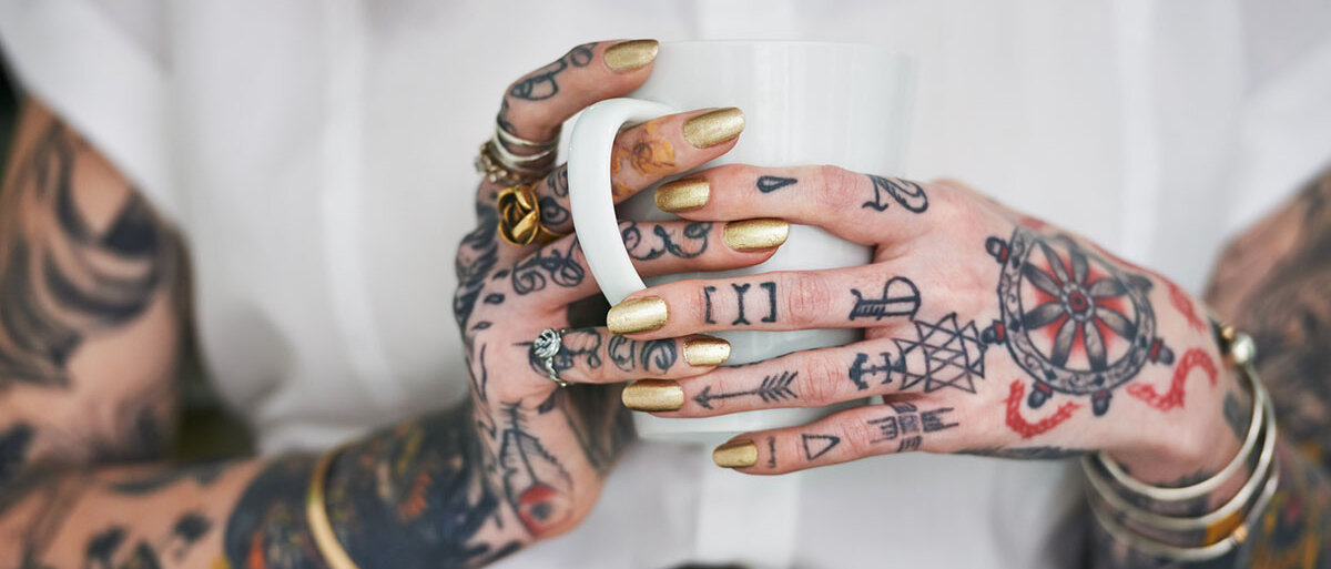 Eine Frau in schwarzem Bleistiftrock und weißer Bluse hält eine Kaffeetasse. Auf den Unterarmen, Handrücken und Fingern hat sie zahlreiche Tattoos. Sie trägt goldenen Nagellack und auffälligen Schmuck.
