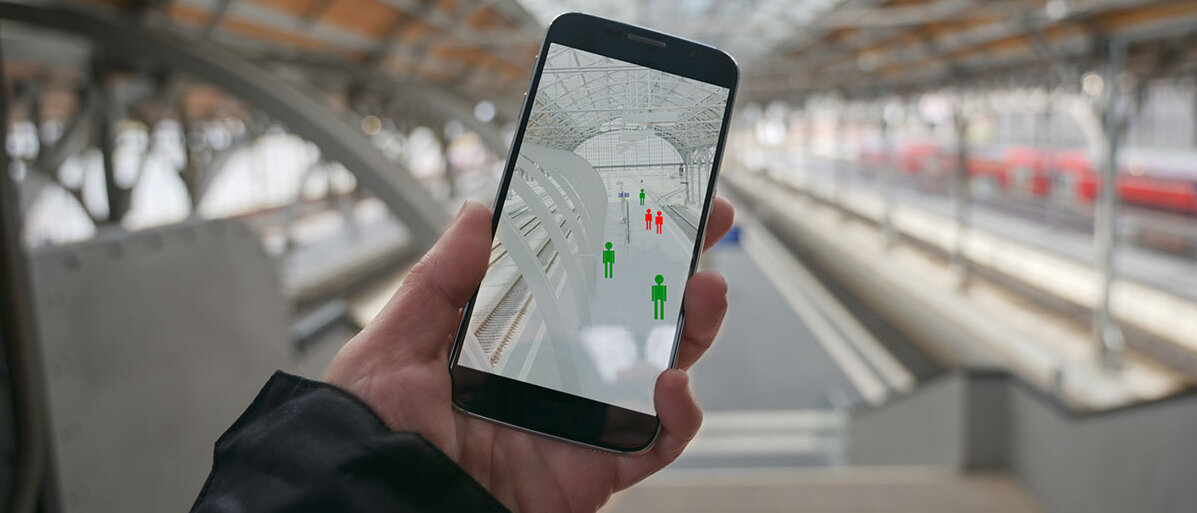 Hand mit einer Smartphone-App zur Nachverfolgung von Infektionsketten; im Hintergrund ein Bahnhof.