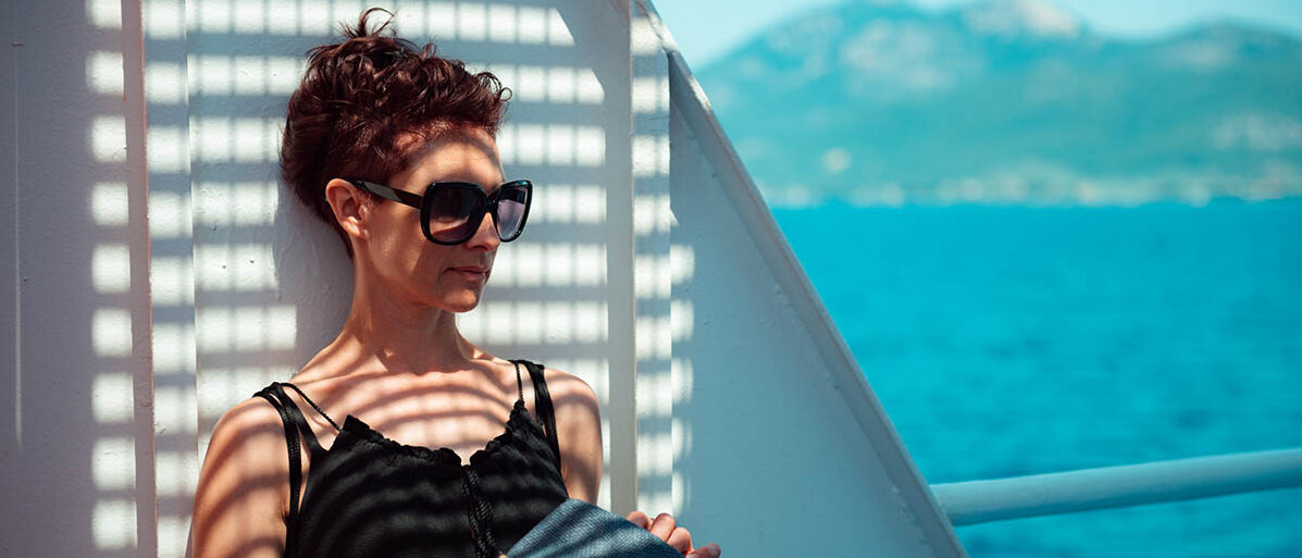 Eine Frau steht auf einer Fähre im Schatten, trägt eine Sonnenbrille und hält einen Strohhut in der Hand.