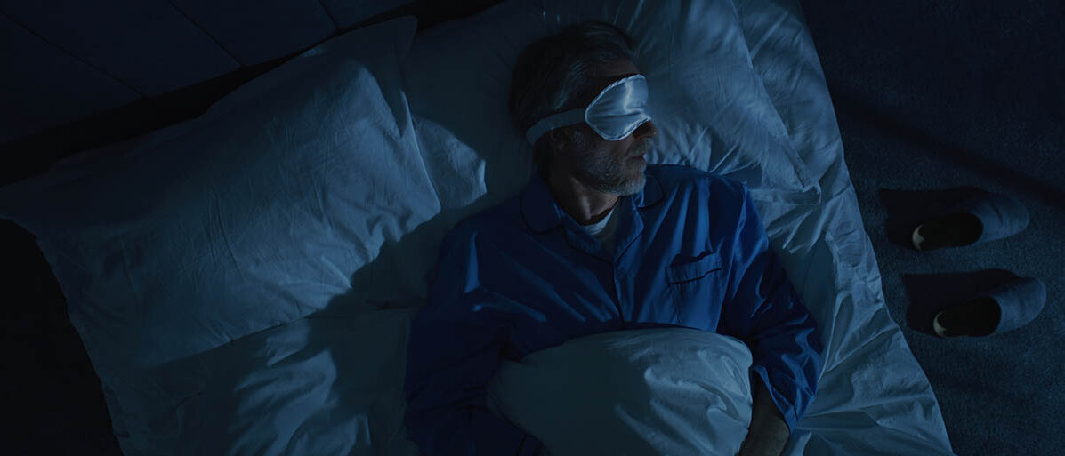 Ein Mann liegt mit Schlafbrille im dunklen Schlafzimmer im Bett.