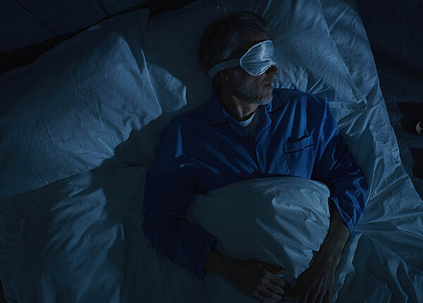 Ein Mann liegt mit Schlafbrille im dunklen Schlafzimmer im Bett.