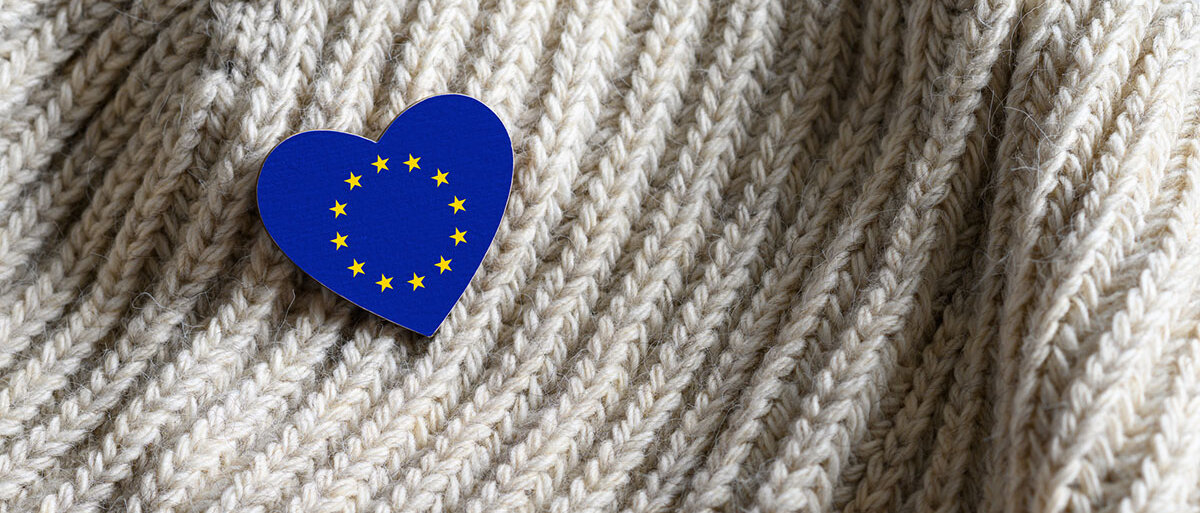 Ein herzförmiger Anstecker zeigt die EU-Flagge. Er steckt in einem Wollpullover oder -schal.
