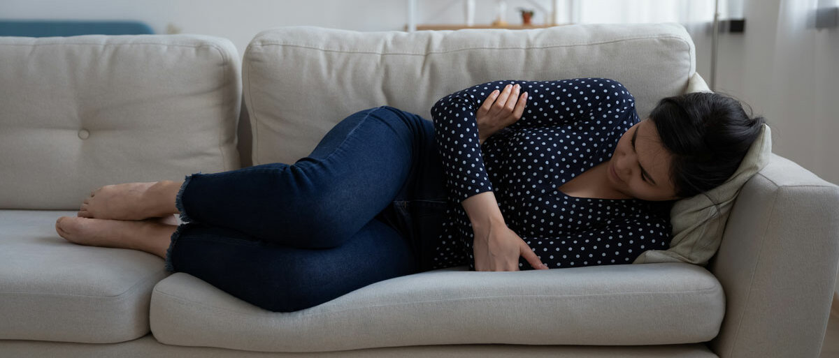 eine Frau liegt mit angezogenen Beinen auf dem Sofa. Sie sieht traurig aus und hat die Arme vor dem Bauch verschränkt.