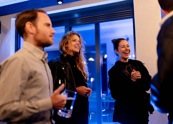 Vier Personen in einer Bar. Ein Mann steht mit dem Rücken zu uns. Zwei Frauen wenden sich ihm zu und lachen. Ein weiterer Mann steht in der Runde und hält einen Bierkrug mit Wasser.