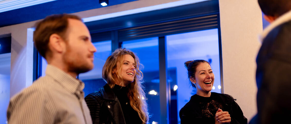 Vier Personen in einer Bar. Ein Mann steht mit dem Rücken zu uns. Zwei Frauen wenden sich ihm zu und lachen. Ein weiterer Mann steht in der Runde und hält einen Bierkrug mit Wasser.