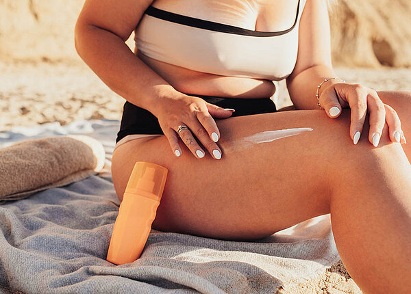 Eine Frau sitzt am Strand auf ihrem Handtuch. Sie cremt sich den Oberschenkel ein, an ihr Bein ist eine Flasche Sonnencreme gelehnt.