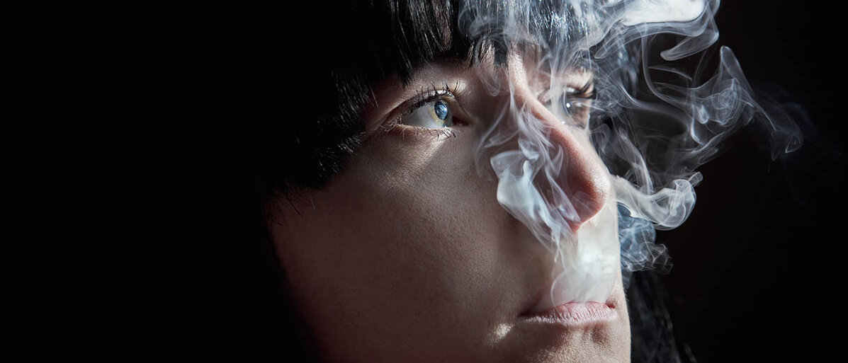 Eine Frau bläst aus dem Mund Zigarettenrauch aus, nach oben, an den Augen vorbei.