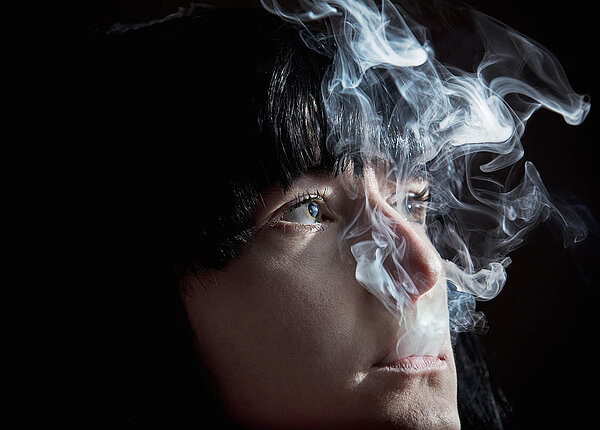 Eine Frau bläst aus dem Mund Zigarettenrauch aus, nach oben, an den Augen vorbei.