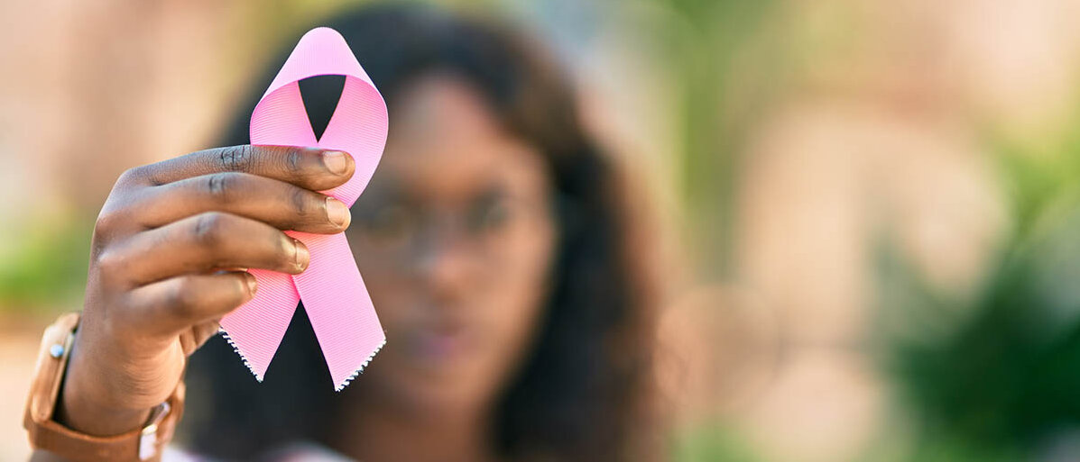 Eine Frau streckt dem Betrachter eine rosa Brustkrebs-Awareness-Schleife entgegen. Die Schleife ist im Fokus, die Frau im Hintergrund unscharf.