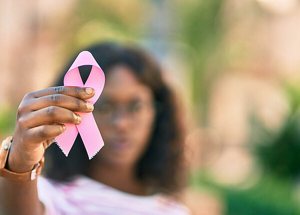 Eine Frau streckt dem Betrachter eine rosa Brustkrebs-Awareness-Schleife entgegen. Die Schleife ist im Fokus, die Frau im Hintergrund unscharf.