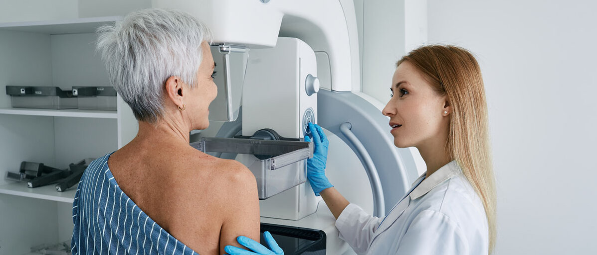 Eine Frau mit kurzen grauen Haaren steht vor einem Untersuchungsgerät zur Mammographie. Eine Ärztin hält sie am Arm, lächelt und stellt das Gerät ein.