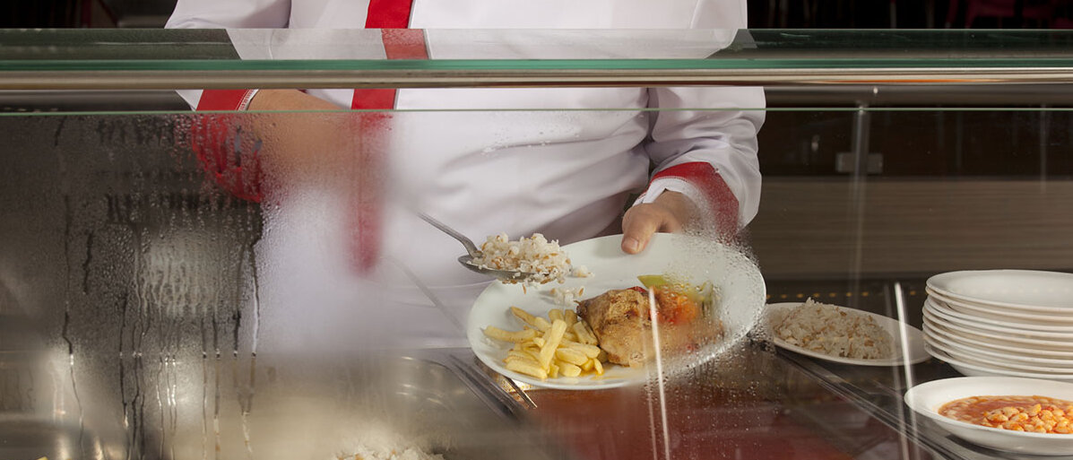 Ein Koch füllt hinter einer Plexiglastrennwand einen Teller mit Fleisch und Pommes.