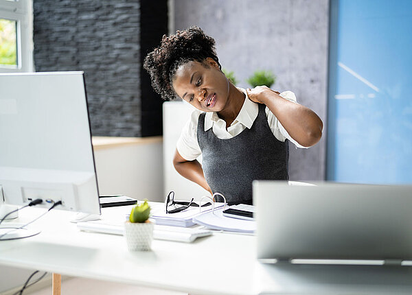 Schwarze Frau greift sich an ihrem Computerarbeitsplatz in den Nacken.