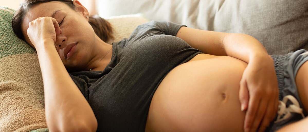 Eine schwangere Frau liegt auf dem Sofa, verzieht das Gesicht und fasst sich an die Stirn.
