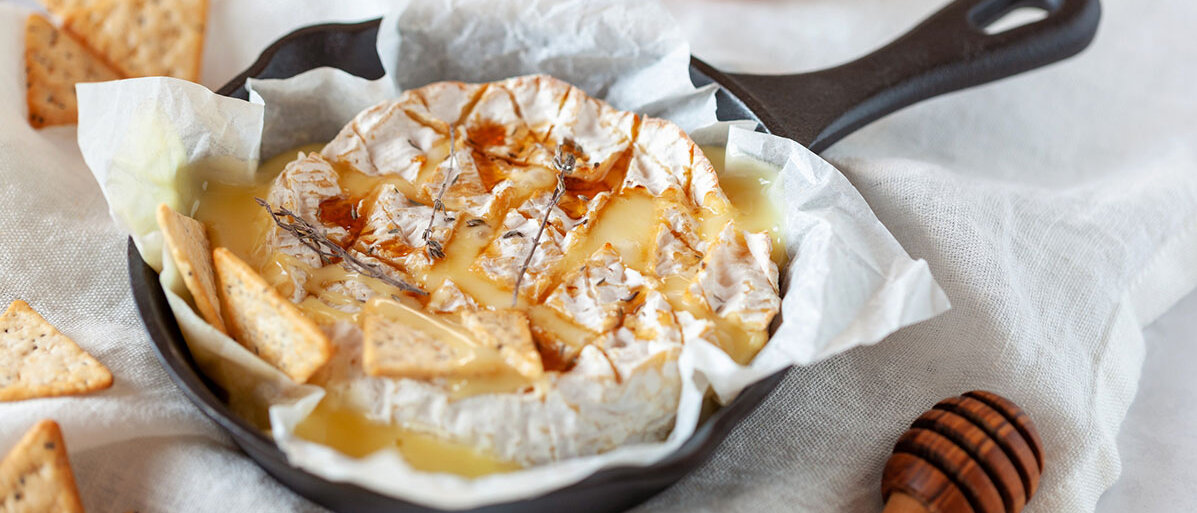 Ofenkaese mit einer weißen Serviette in einer schwarzen Gusspfanne. Drum herum liegen Kraecker und ein Honigloeffel