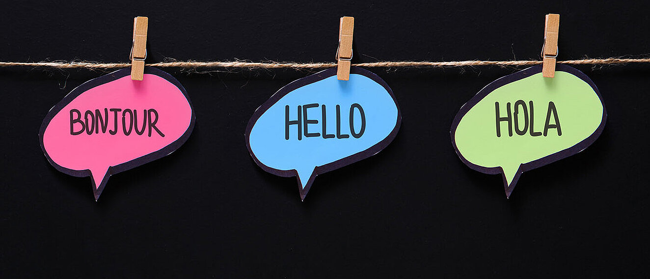 Mehrsprachigkeit schützt das Gehirn drei unterschiedliche bunte Sprechblasen mit verschiedenen Sprachen aufgehängt an einem querverlaufenden Faden mit Holzklammern