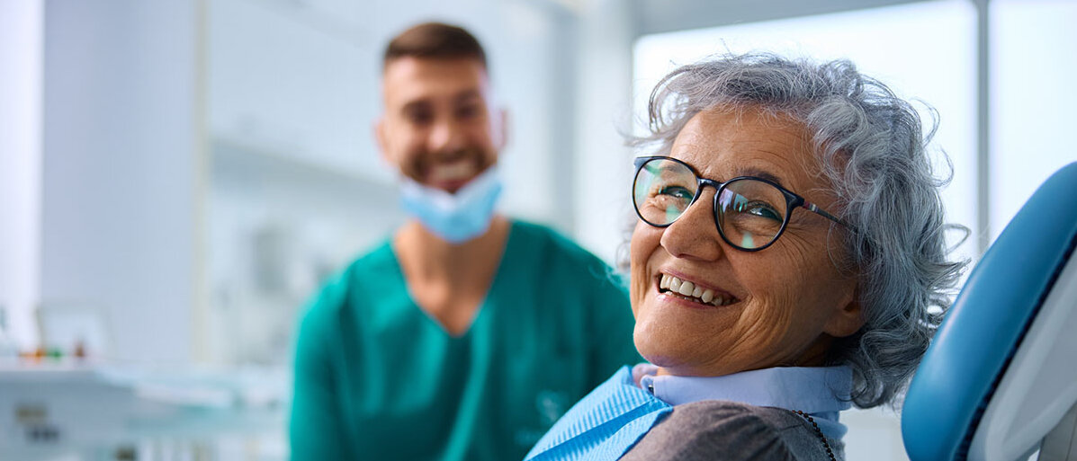 fröhliche Seniorin im Zahnarztstuhl blickt direkt in die Kamera; im Hintergrund ein lächelnder Zahnarzt 