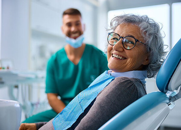 fröhliche Seniorin im Zahnarztstuhl blickt direkt in die Kamera; im Hintergrund ein lächelnder Zahnarzt 
