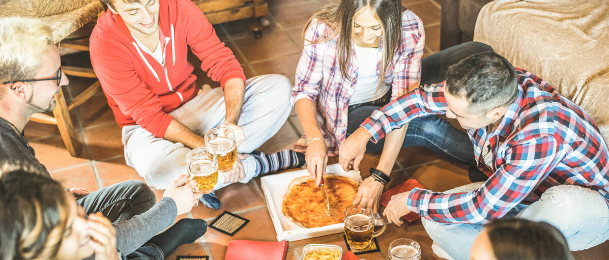 Sechs Junge Menschen sitzen auf dem Fußboden, essen Pizza und Pommes und trinken Bier.
