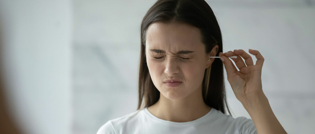 Eine junge Frau reinigt sich das Ohr mit einem Wattestäbchen. Sie verkneift dabei ihr Gesicht.