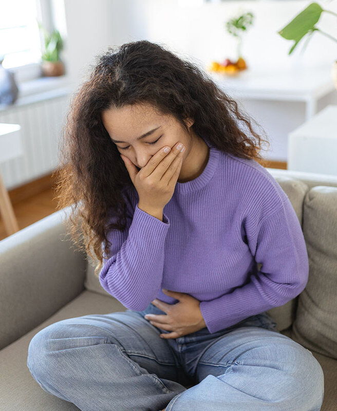 Eine Frau sitzt gekrümmt, hält sich mit einer Hand den Bauch, die andere hält sie vor den Mund.