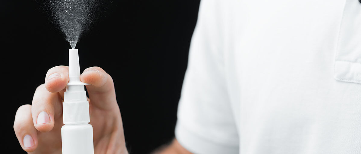 Mann in weißem Hemd mit Nasenspray in der Hand. Er gibt einen Sprühstrahl ab vor schwarzem Hintergrund.