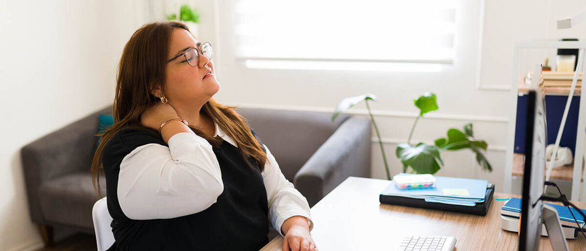 Eine Frau sitzt am Schreibtisch vor dem Computer. Sie hat den Kopf in den Nacken gelegt und hält sich den Nacken, dabei ist ihr Gesicht schmerzverzerrt.
