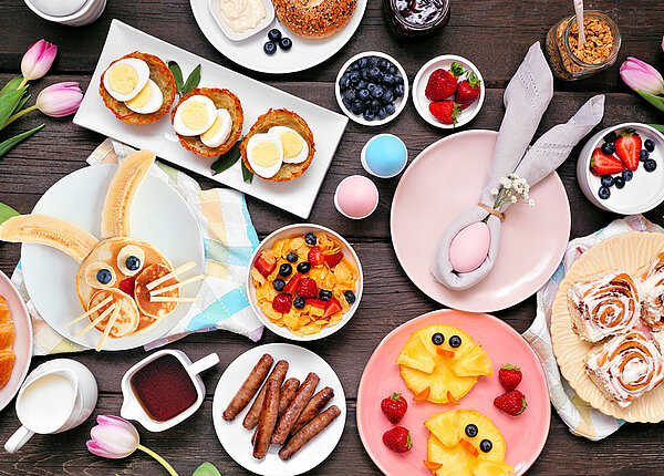 Blick von oben auf einen Ostertisch: Zwischen Tulpen und bunten Ostereiern stehen Teller mit verschiedenen Speisen, zum Beispiel einem Osterhasengesicht aus Pancakes oder einem Küken aus Ananas.