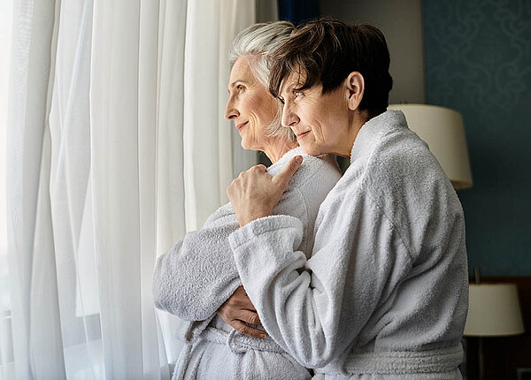 Zwei Seniorinnen im Bademantel stehen am Fenster und kuscheln.