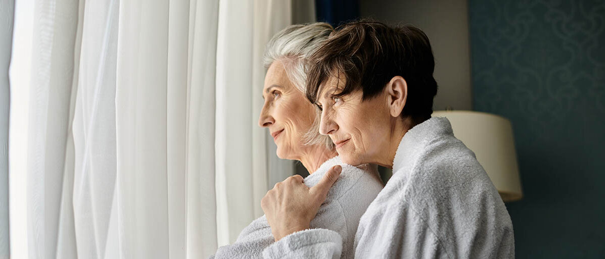 Zwei Seniorinnen im Bademantel stehen am Fenster und kuscheln.