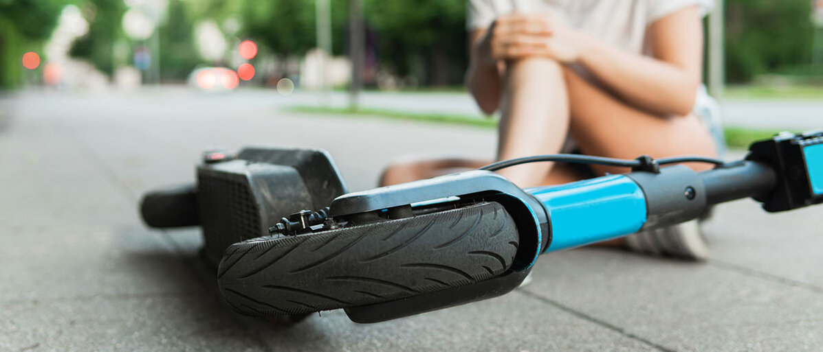 Frau hatte E-Scooter-Unfall und sitzt jetzt vor dem Gefährt auf der Srtraße. Sie hält sich das Bein.