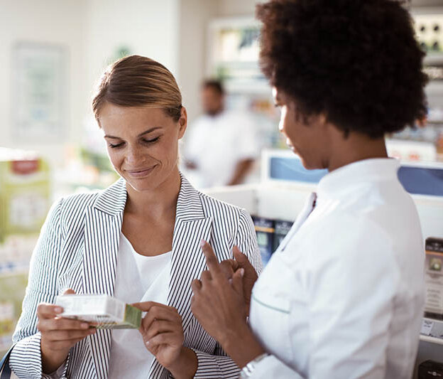 Eine Apothekenkundin hält ein Arzneimittel in der Hand. Die PTA oder Apothekerin erklärt ihr etwas. Die Kundin lächelt.
