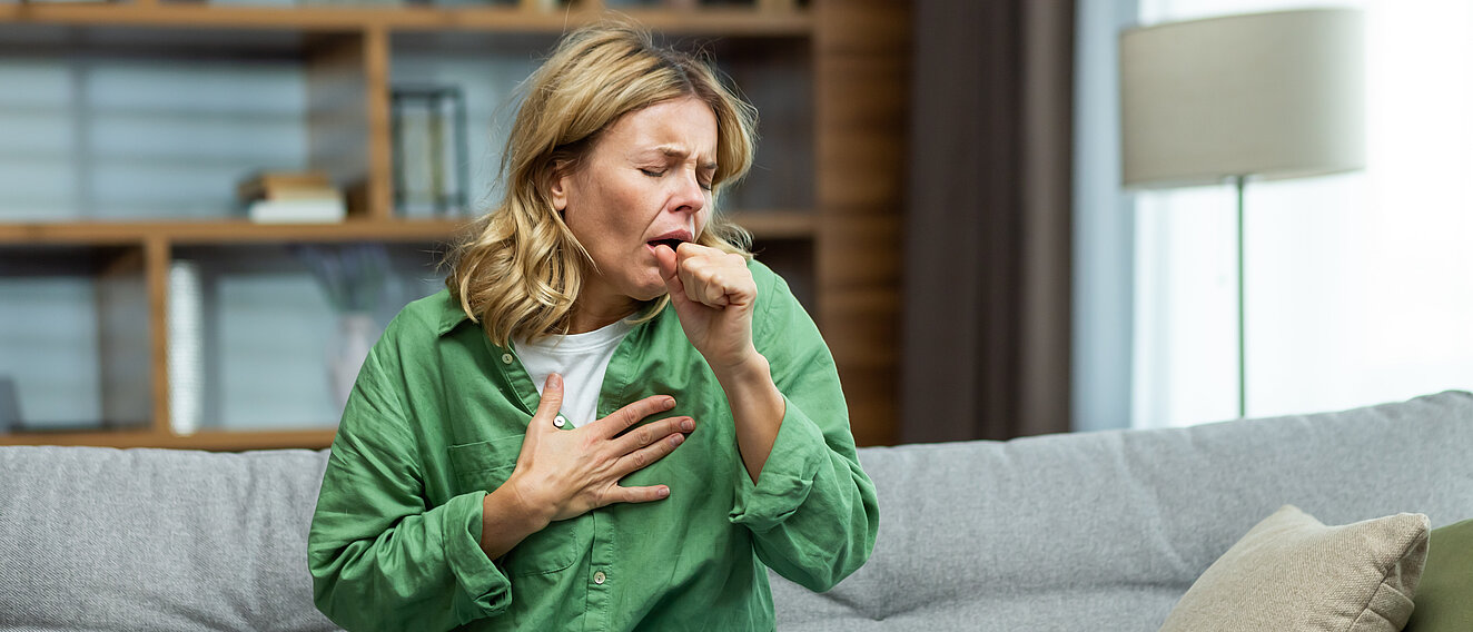 Auf der Couch sitzt eine Frau, die heftig in ihre zur Faust geformte Hand hustet. Ihre zweite Hand liegt flach auf der Brust, ihr Gesicht zeigt deutliche Schmerzen.