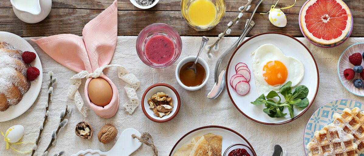 Auf dem Tisch stehen Kuchen, Waffeln, ein Osterei, das in die Serviette eingeschlagen wie ein Hase aussieht und viele weitere Frühstücks-Leckereien.