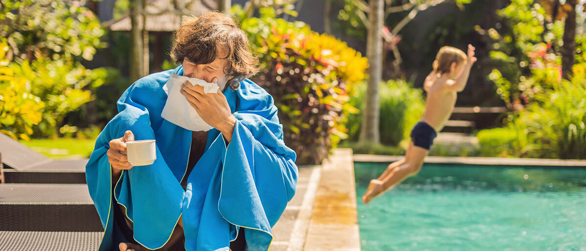 Leisure Sickness: Im Urlaub krank durch Stress Ein Mann sitzt eingewickelt in eine Decke auf einer Sonnenliege am Pool. Er schnäuzt sich die Nase und hält eine Tasse. Die Sonne scheint. Im Hintergrund springt ein Kind in den Pool.