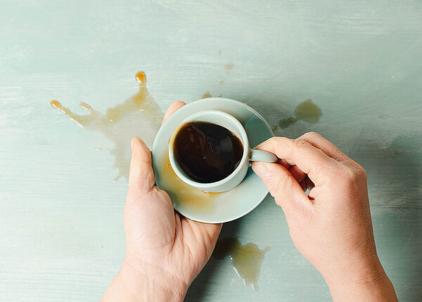 Parkinson: Symptome, Behandlung und Diagnose Blick von oben: Jemand hält eine Kaffeetasse und Untertasse stabilisierend mit beiden Händen, doch viel Kaffee wurde verschüttet.