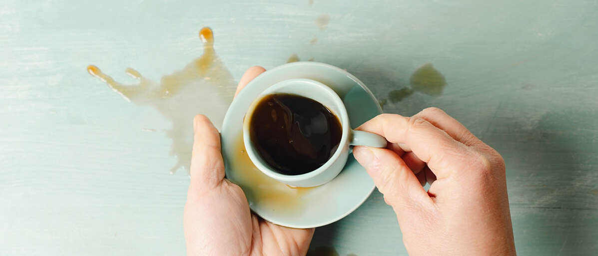 Parkinson: Symptome, Behandlung und Diagnose Blick von oben: Jemand hält eine Kaffeetasse und Untertasse stabilisierend mit beiden Händen, doch viel Kaffee wurde verschüttet.