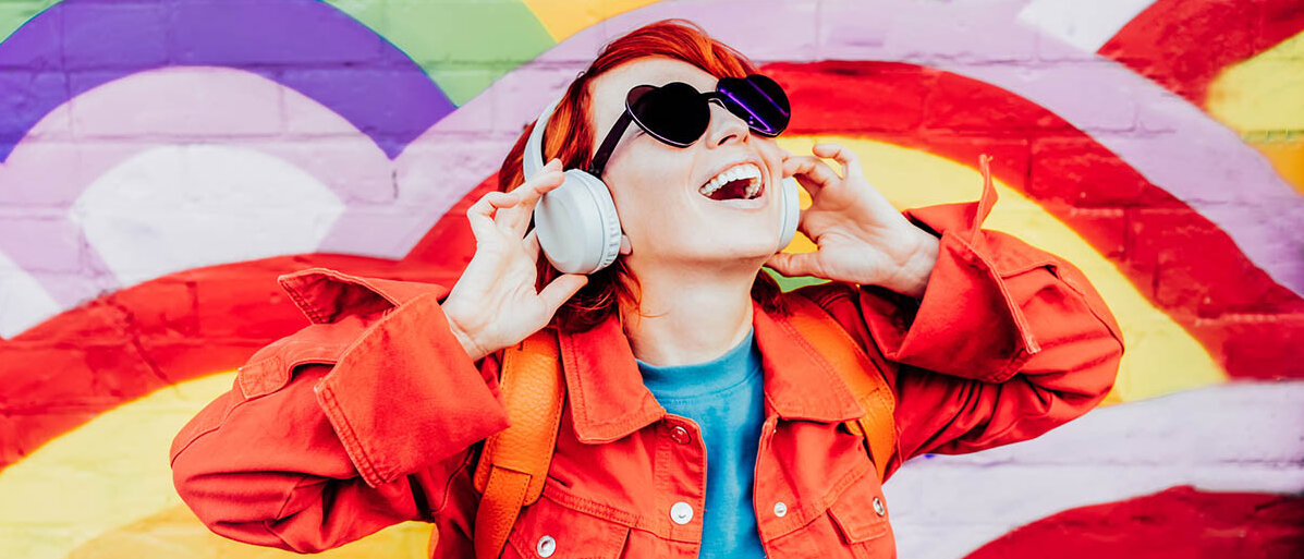 Synästhesie-Wahrnehmungen betreffen alle Sinne. Es gibt verschiedene Formen, zum Beispiel die Graphem-Farb-Anästhesie. Eine junge Frau hört Musik über ihre Kopfhörer. Sie ist bunt gekleidet, hat knallrote Haare und die Mauer hinter ihr ist mit Regenbögen bemalt.