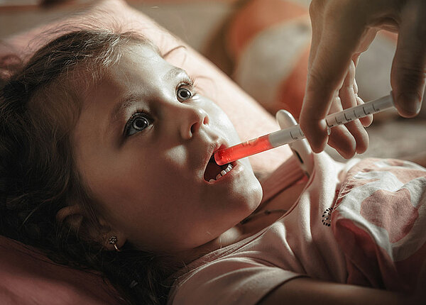 Ein Mädchen im Kindergartenalter liegt im Bett und bekommt eine pinke Suspension aus einer Dosierspritze in den Mund verabreicht.