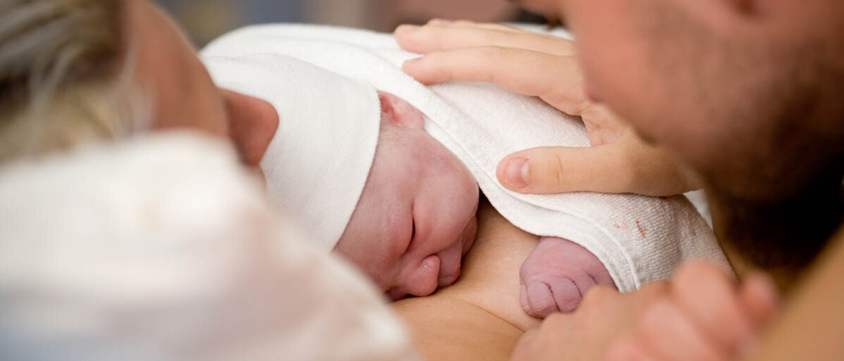 Bonding ist der Begriff für Hautkontakt durch etwa Kuscheln nach der Geburt. Im Krankenhausbett liegt ein Neugeborenes auf dem Bauch der Mutter, der Vater streichelt es.