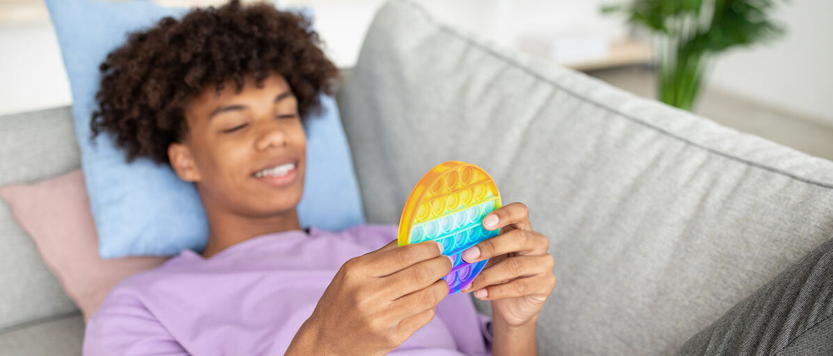 Macht ADHS kreativer? Ein Teenager Junge liegt auf einem Sofa und spielt mit einem bunten Fidget Toy.