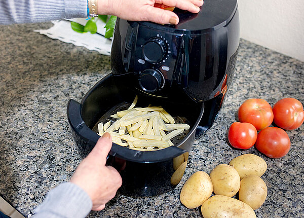 Ein geöffneter Airfryer, in dessen Schubfach Pommes sind. Nebendran liegen Tomaten und Kartoffel