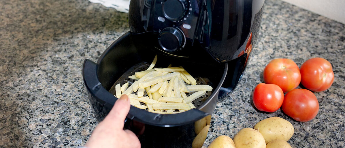 Ein geöffneter Airfryer, in dessen Schubfach Pommes sind. Nebendran liegen Tomaten und Kartoffel