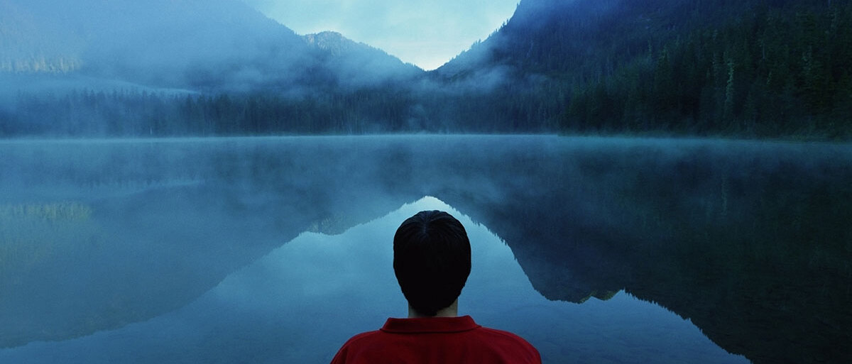 Ein Mann steht mit dem Rücken zu uns vor einem nebelverhangenen Bergsee.